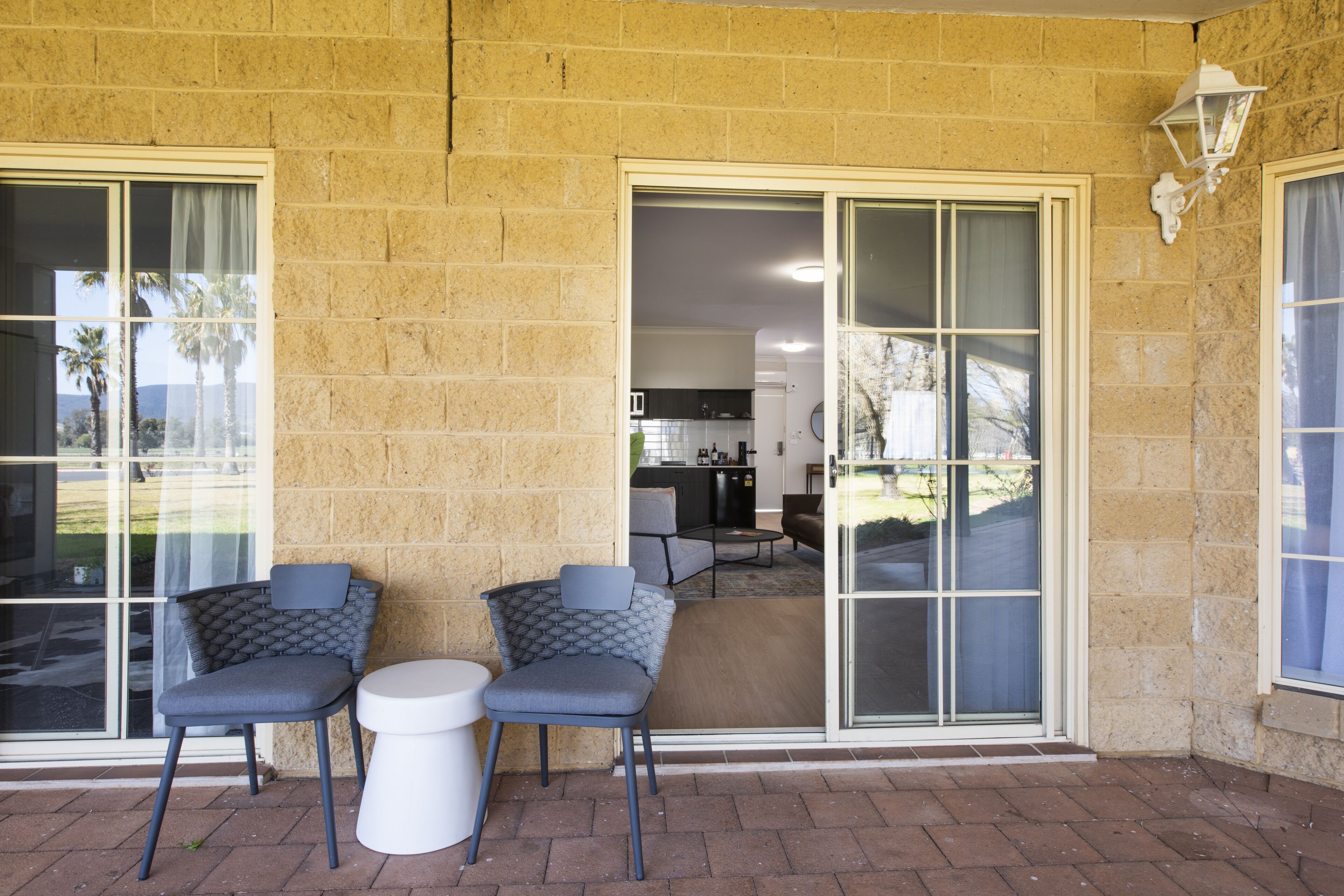 A patio with two chairs and a small table, a sliding glass door leading to a kitchen, and a brick wall.