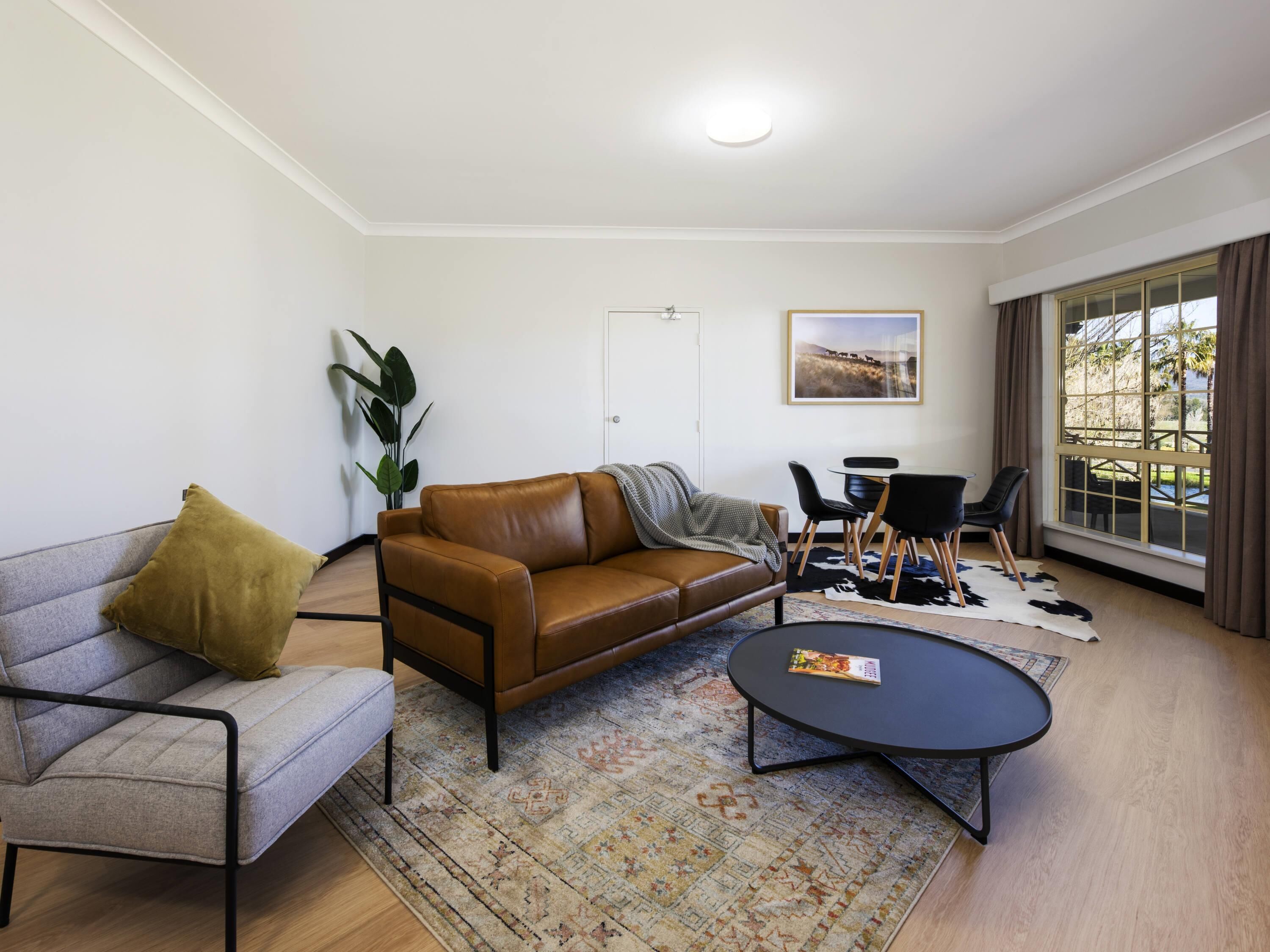 A living room with a brown leather sofa, a grey armchair, a black coffee table, a dining table with chairs, and a large window with curtains.
