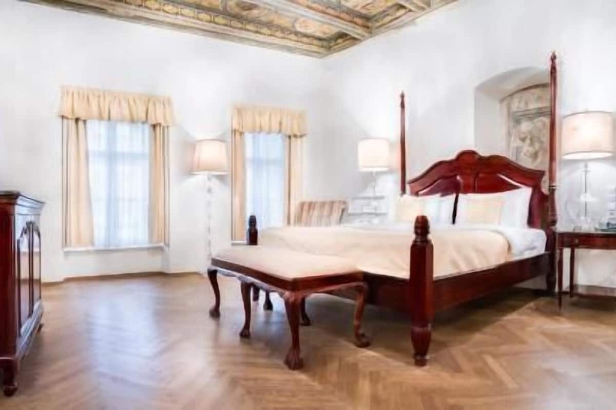 A bedroom with a four-poster bed, a wooden dresser, two bedside lamps, and a ceiling with decorative plasterwork.