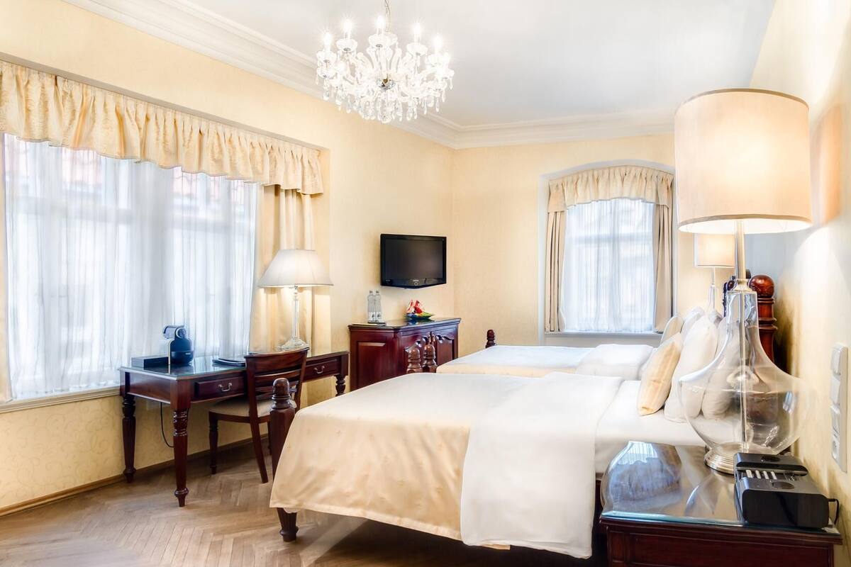 A hotel room with two beds, a desk with a lamp, a TV, and a chandelier.