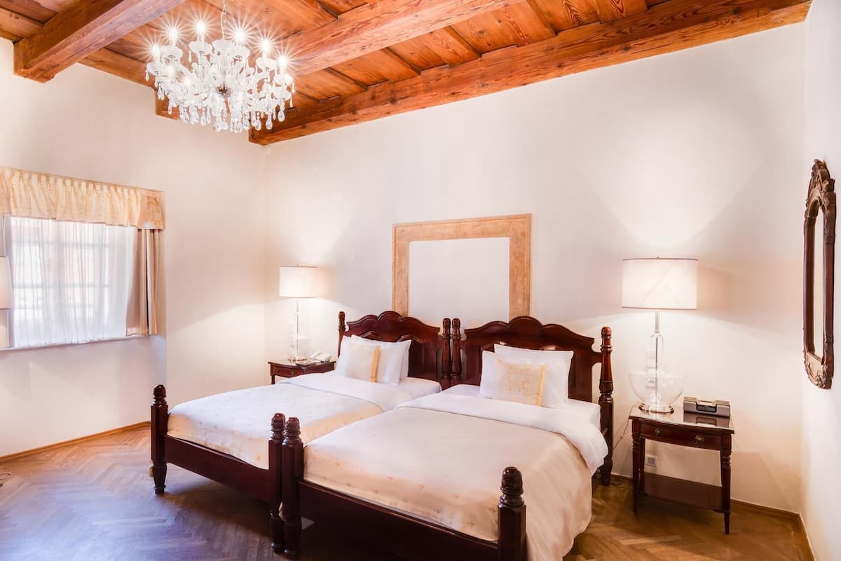 A double bed with a wooden headboard, two bedside tables with lamps, a chandelier, and a window with curtains.