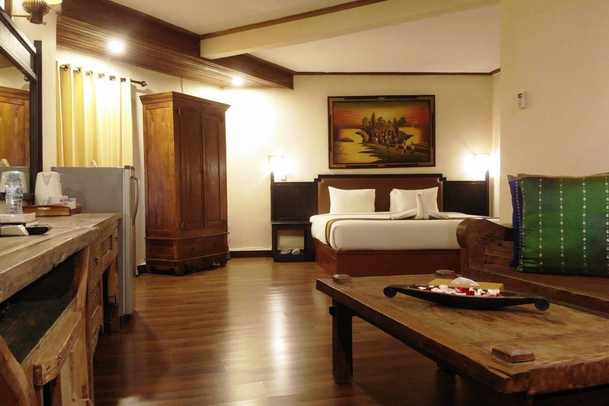 A hotel room with a wooden desk, a bed with white bedding, a painting on the wall, and a green pillow on the couch.
