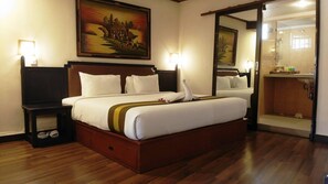 Habitación de hotel con una cama grande, cabecera de madera, un cuadro sobre la cama, un espejo y un baño visible a través de una puerta abierta.