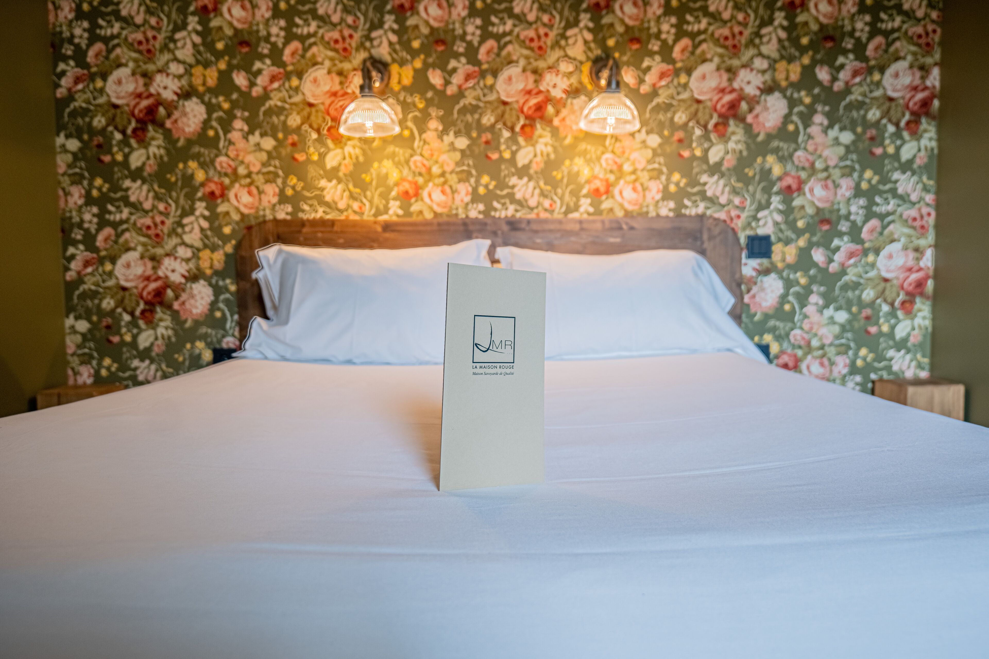 Een net opgemaakt bed met witte lakens en een wit kussen, voor een behang met bloemen.