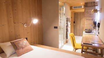 A bedroom with a wooden ceiling, a bed with a white and pink pillow, a bedside table with a remote control, a yellow chair, and a bathroom visible through an open door.