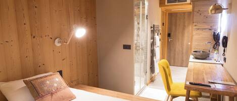 A bedroom with a wooden ceiling, a bed with a white and pink pillow, a bedside table with a remote control, a yellow chair, and a bathroom visible through an open door.