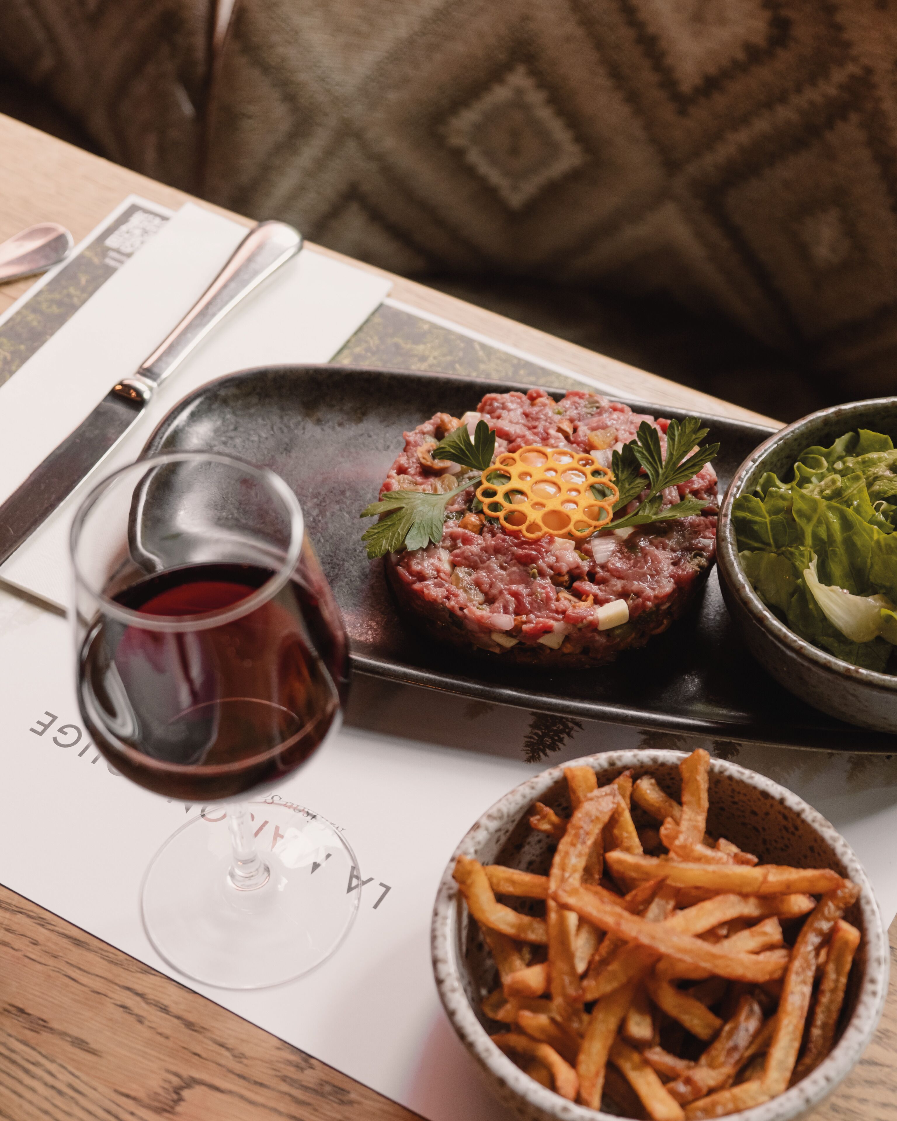 A dining table with a steak tartare dish, a glass of red wine, and a bowl of fries.