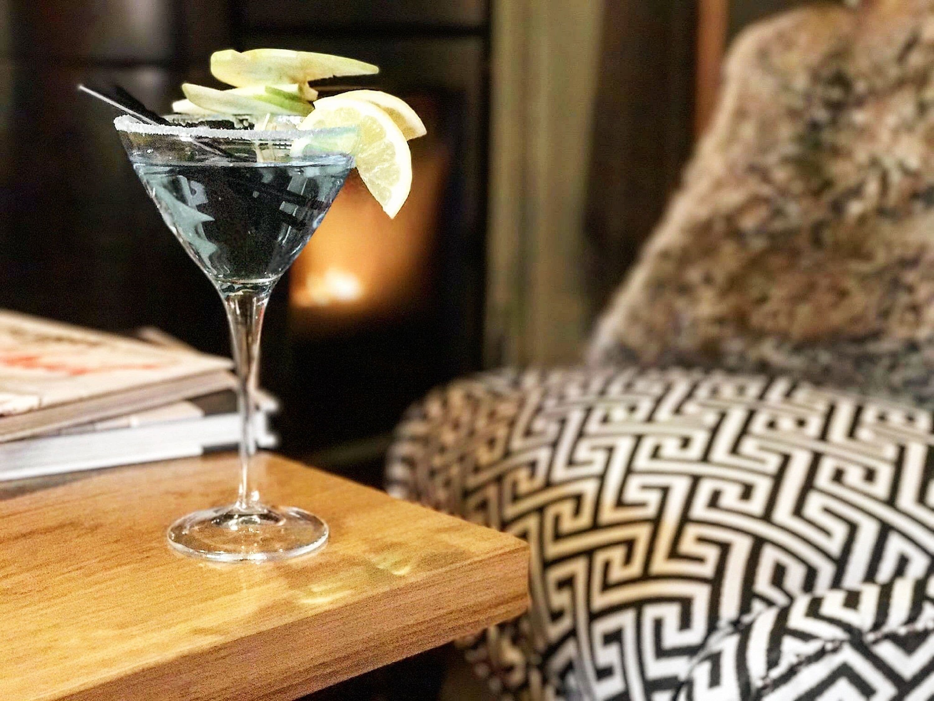 Un cóctel en una copa de martini sobre una mesa de madera, con un cojín estampado al fondo.