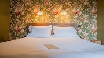 Una cama bien tendida con sábanas blancas y una tarjeta sobre la cama. La habitación tiene un papel tapiz floral y dos lámparas fijadas a la pared.
