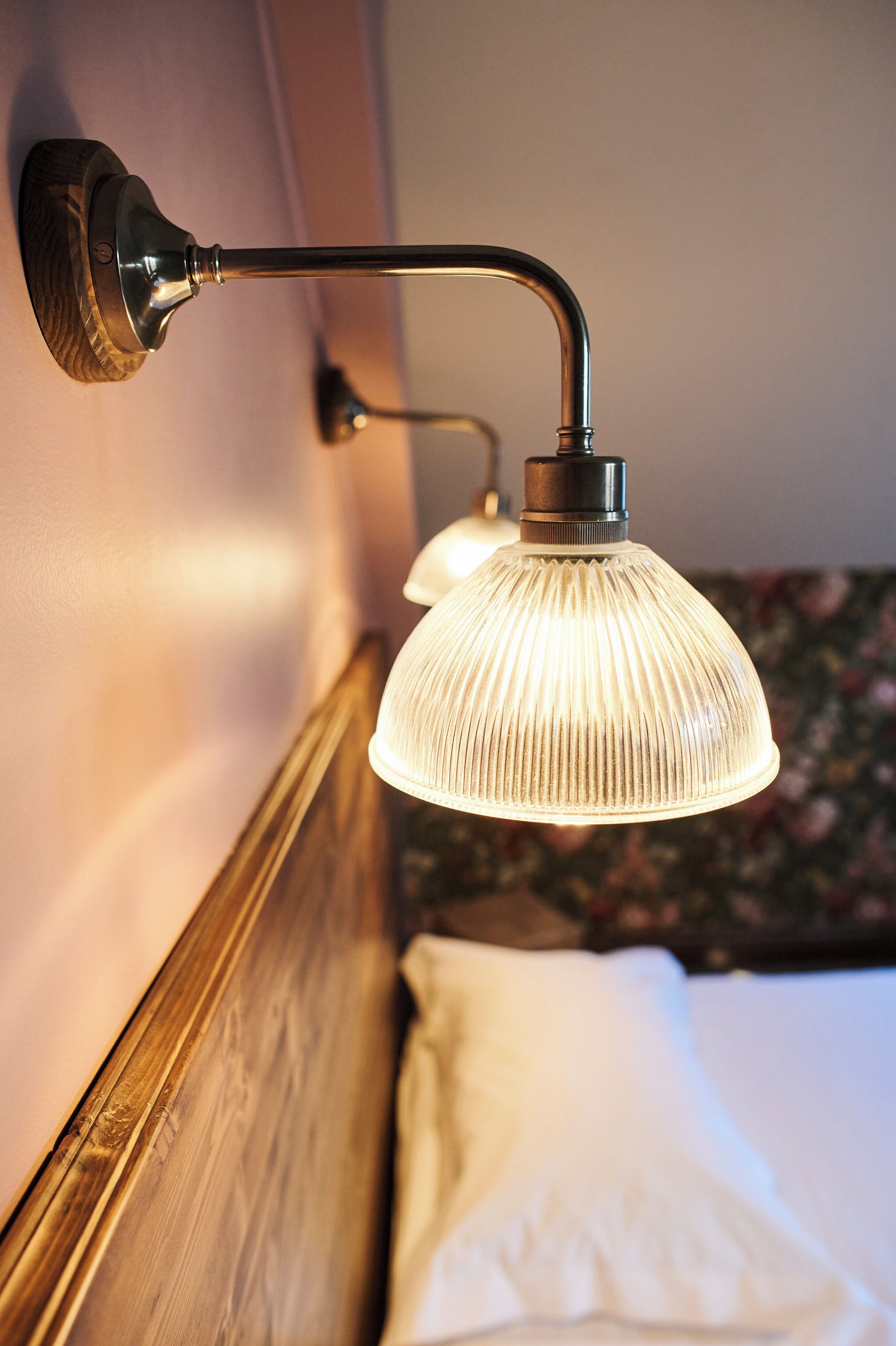 Een muurlamp met een gebogen arm en een gegroefde glazen kap, boven een houten hoofdbord en een wit kussen.