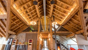 A spacious loft-style room with exposed wooden beams, a central staircase, a fireplace, and a kitchen area with a refrigerator and shelves. - La Maison Rouge (Barberaz)