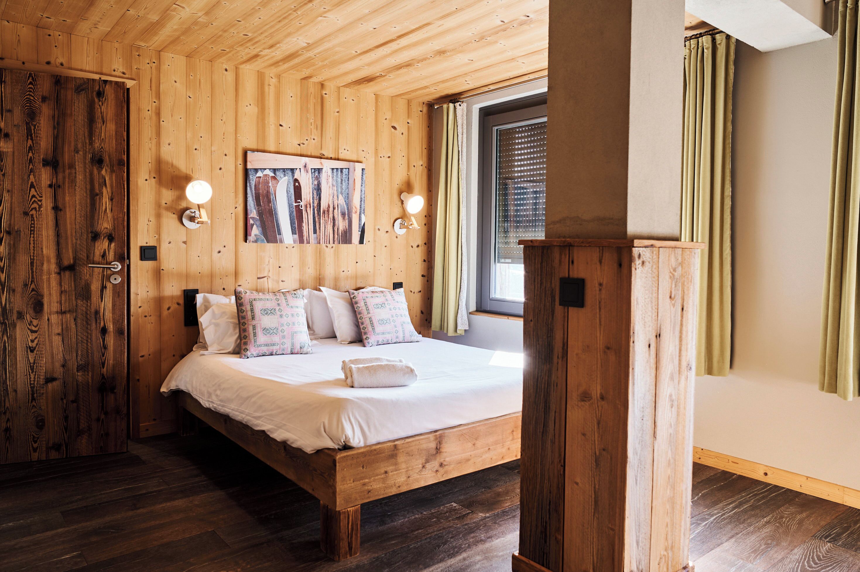 A wooden-paneled bedroom with a bed, two wall-mounted lights, a window with curtains, and a painting on the wall.