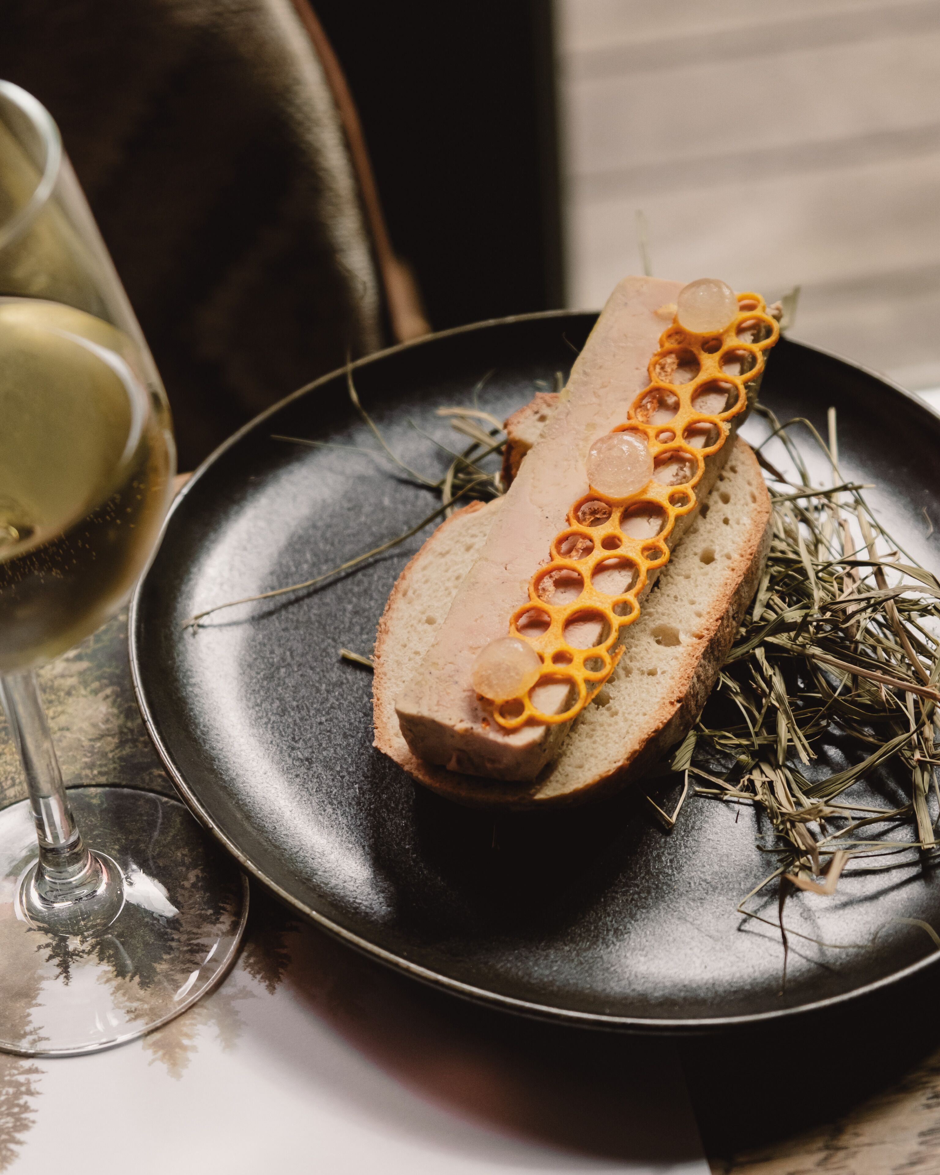 A slice of bread with a decorative topping on a plate, accompanied by a glass of white wine and some herbs.