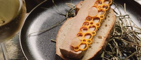 Una rebanada de pan con un adorno decorativo en un plato, acompañada de una copa de vino blanco y algunas hierbas.