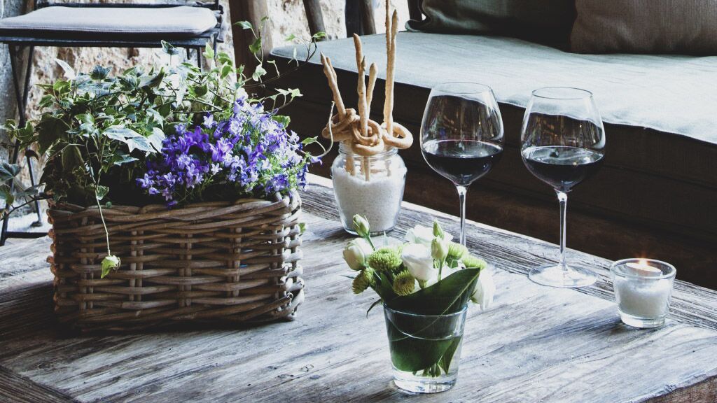 A rustic wooden table with a wicker basket, a glass jar with pretzel sticks, a glass of red wine, a candle, and a small vase with flowers.