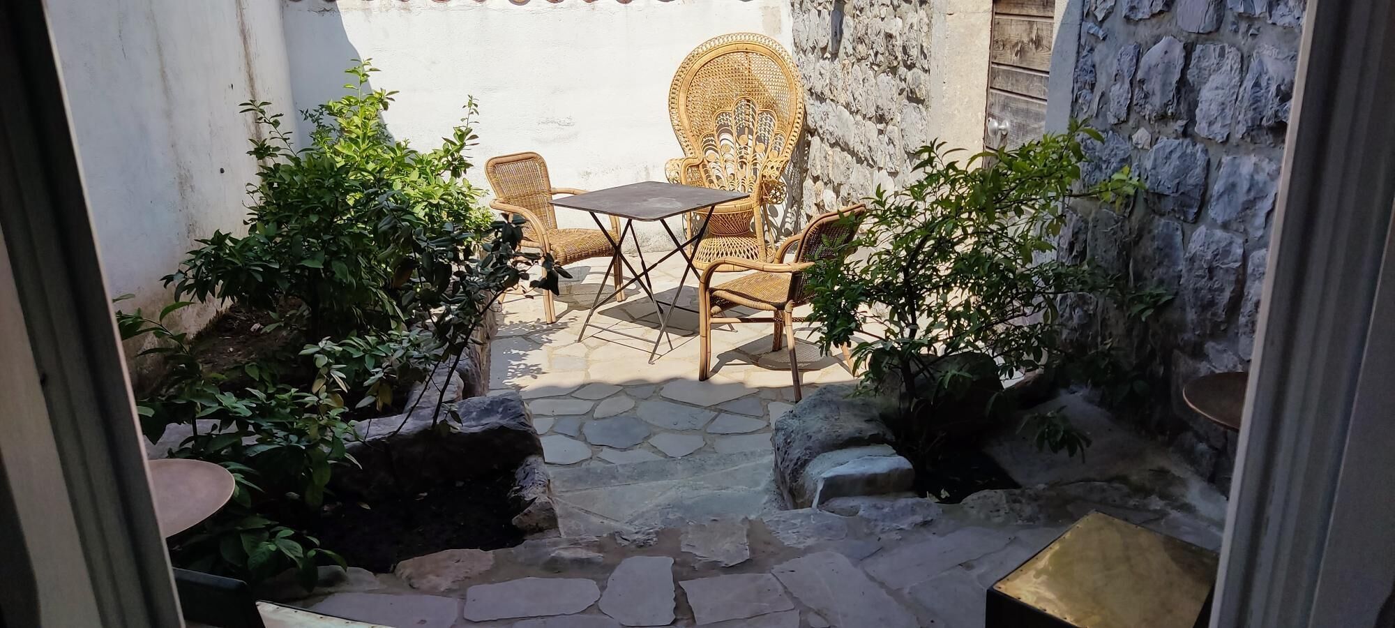Un patio con una mesa de piedra y sillas de mimbre, rodeado de vegetación y muros de piedra.