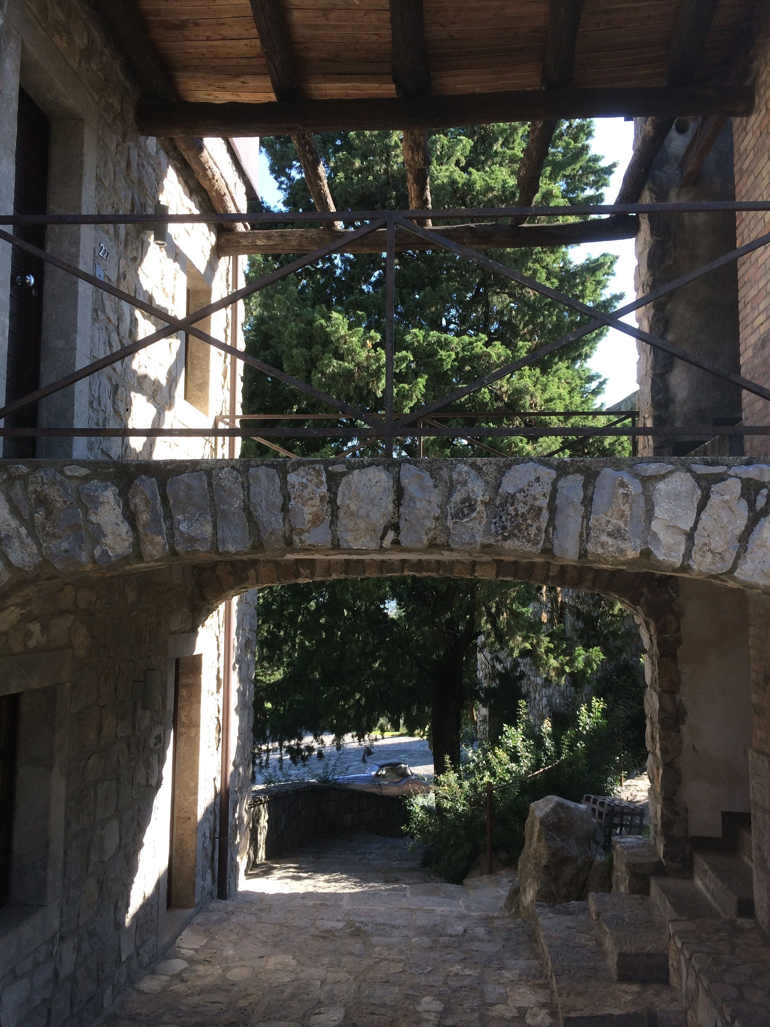 A stone archway leading to a courtyard with a wooden pergola overhead.
