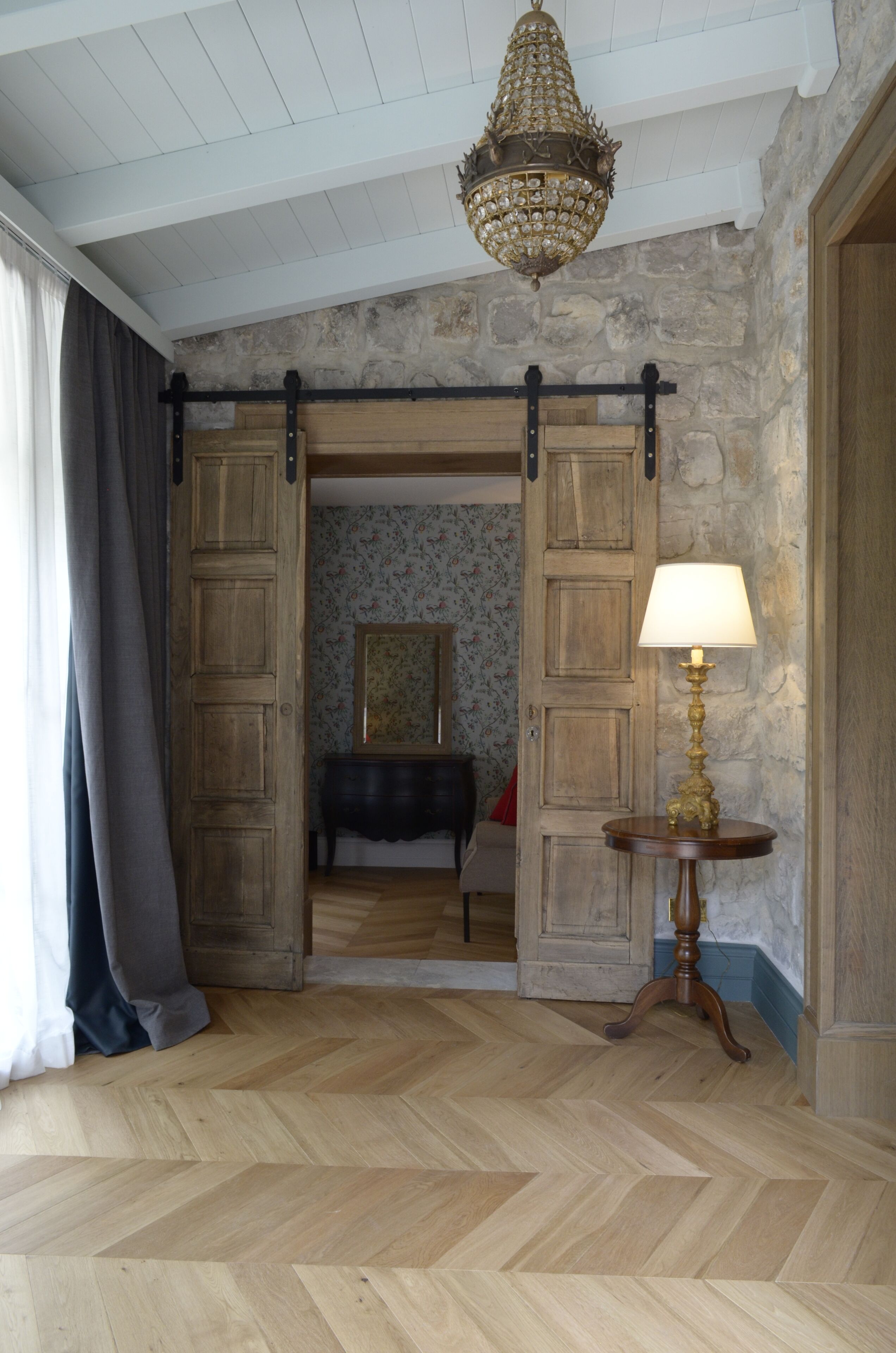 Ein Zimmer mit Fischgrätparkett, einer Chandelüster, einer Holztür mit Spiegel und einem Tisch mit Lampe.