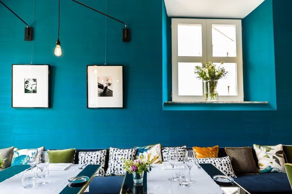 Habitación color turquesa con una mesa de comedor lista para dos personas, cuadros en la pared y una ventana con vistas a zonas verdes.