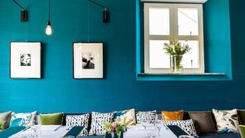 Habitación color turquesa con una mesa de comedor lista para dos personas, cuadros en la pared y una ventana con vistas a zonas verdes.