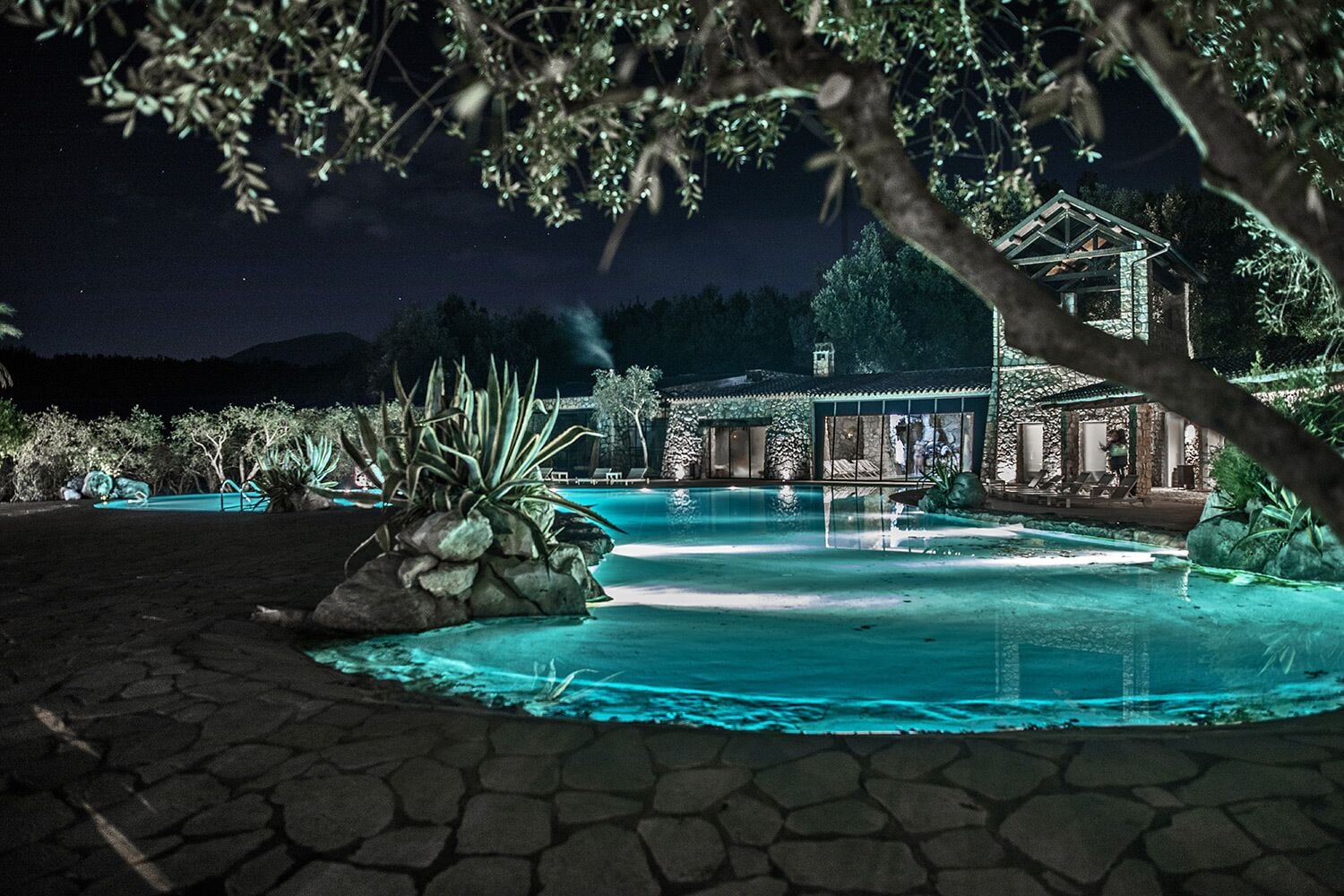 A nighttime view of a resort with a swimming pool, a covered area with lounge chairs, and a building with a stone facade.
