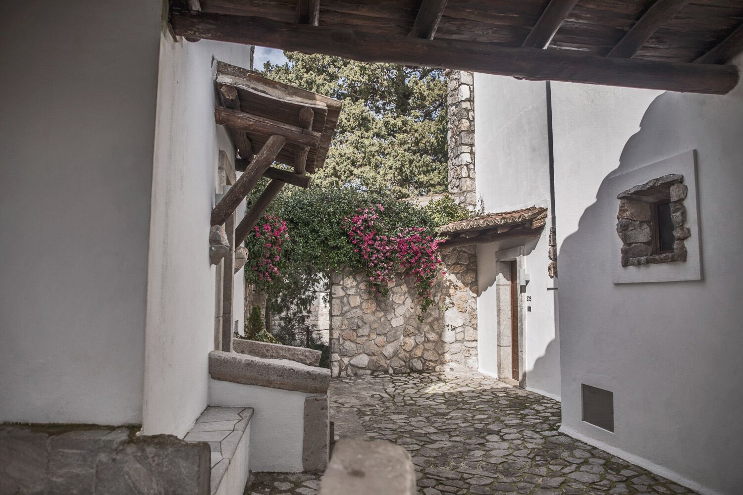 A narrow stone-paved alleyway with a wooden staircase leading to a stone wall with pink flowers.