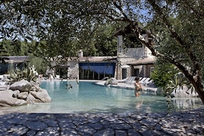 Uma área de piscina com estrutura de pedra, espreguiçadeiras e uma pessoa na água.