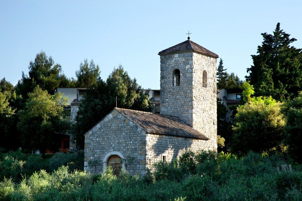 A stone church with a bell tower and a smaller adjacent building, surrounded by greenery.