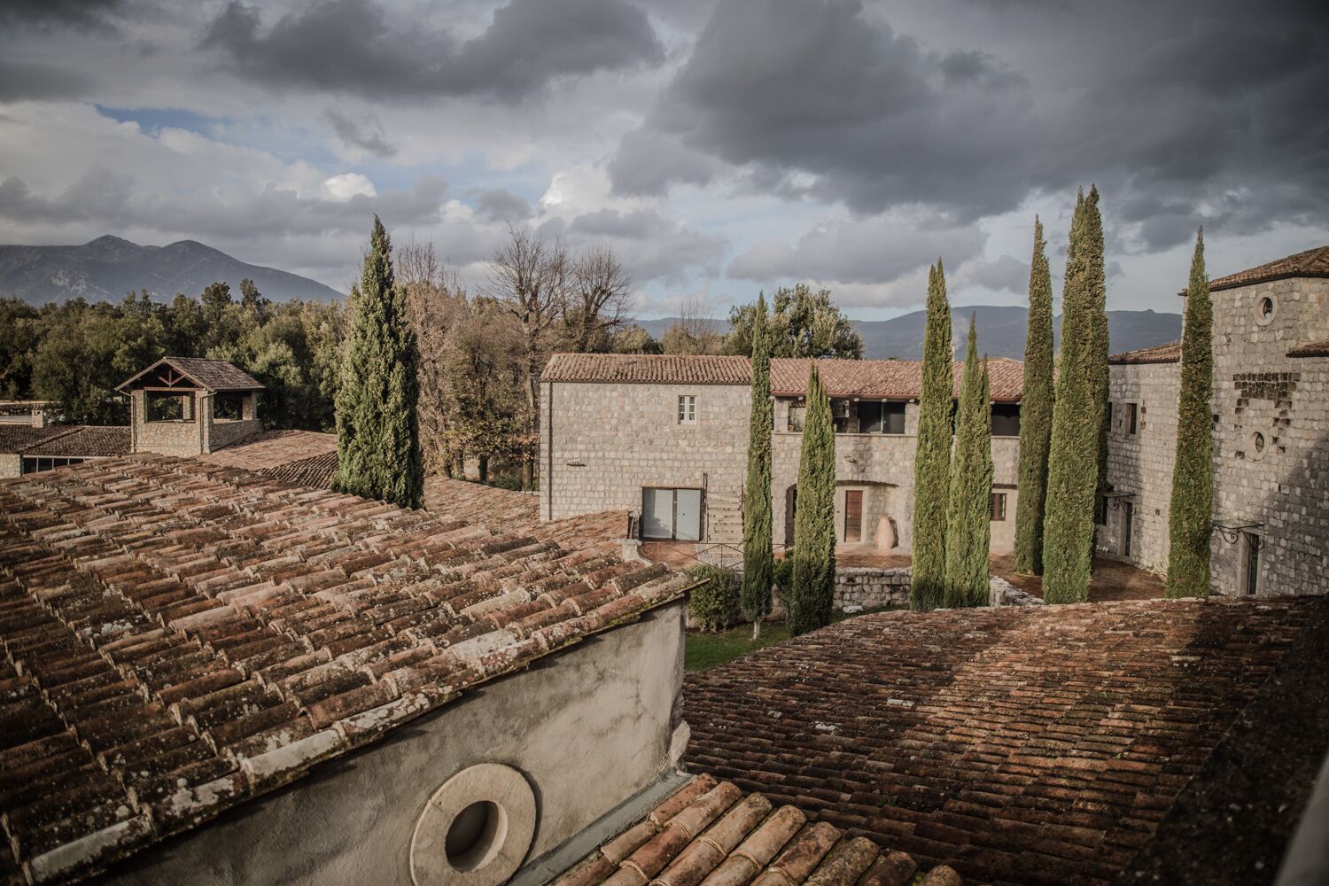 A courtyard with stone buildings, a small wooden structure, and tall cypress trees.