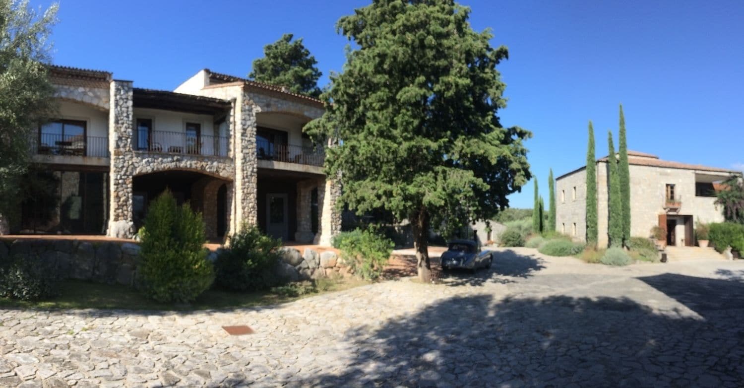 Un bâtiment en pierre doté d’un balcon, entouré de verdure et bordé d’une allée pavée.