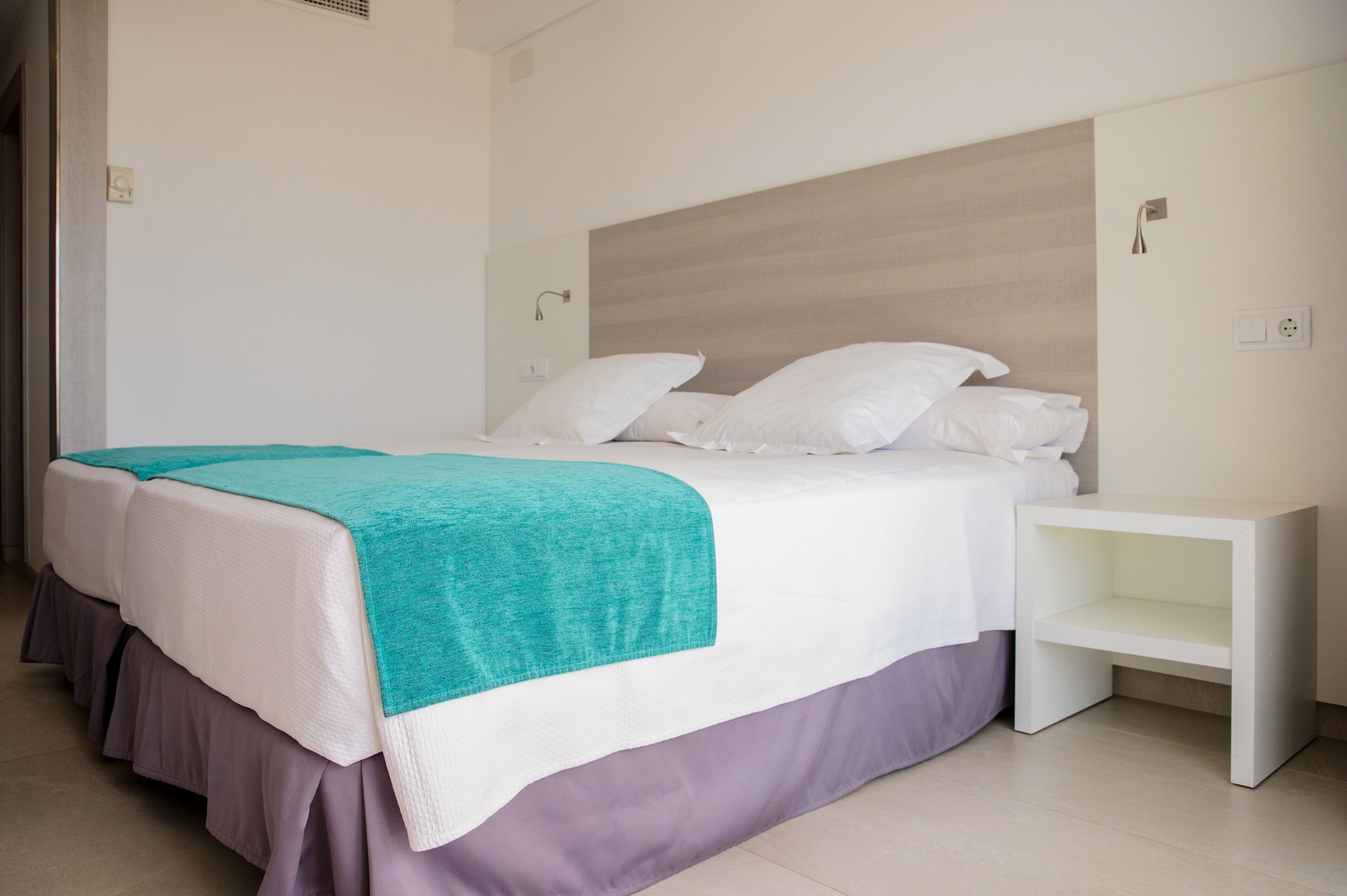 Un lit bien fait, recouvert d’une couverture turquoise, avec une tête de lit blanche et une table de chevet blanche à côté.
