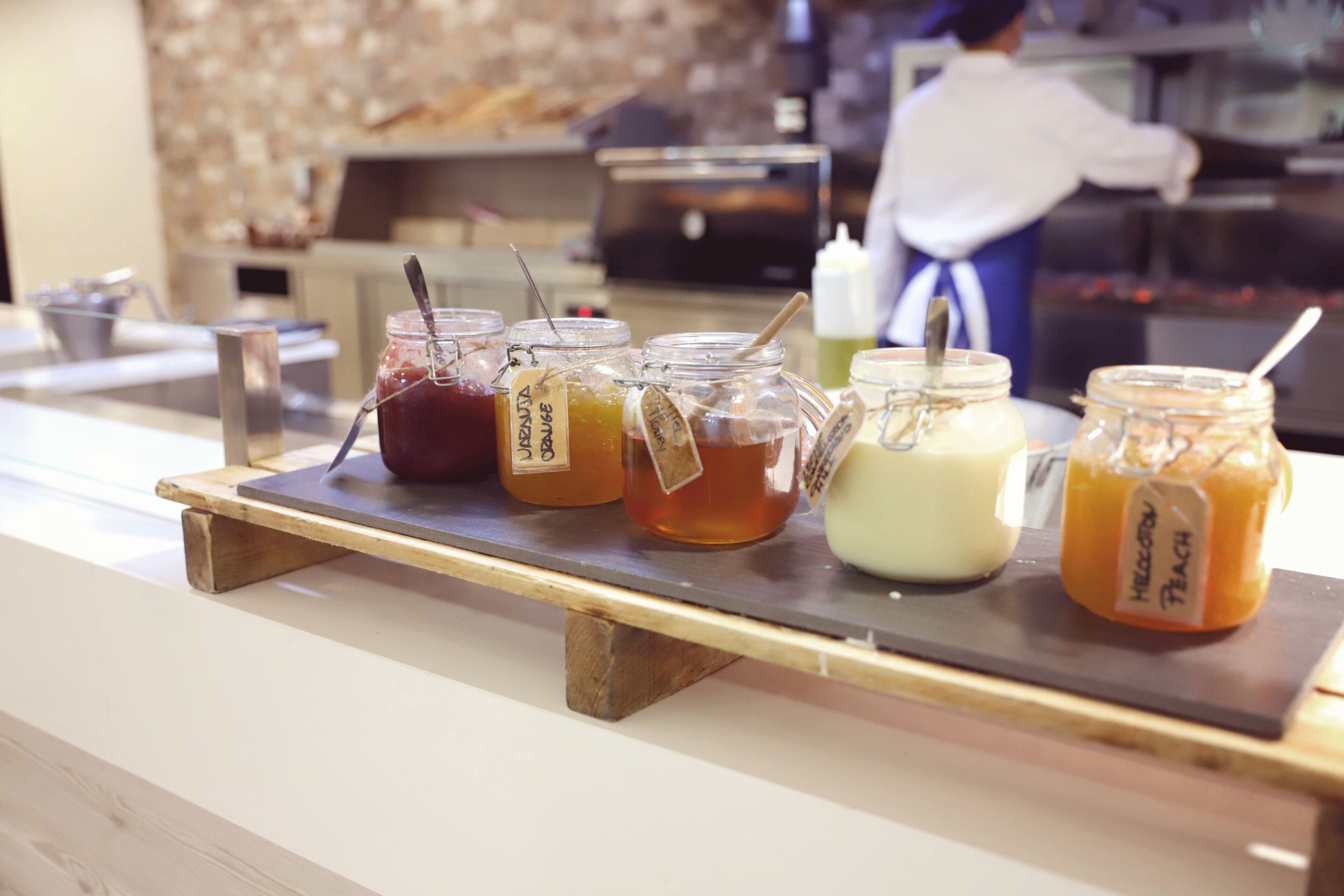 A selection of jars with labels on a wooden board, likely containing condiments or preserves, in a commercial setting.