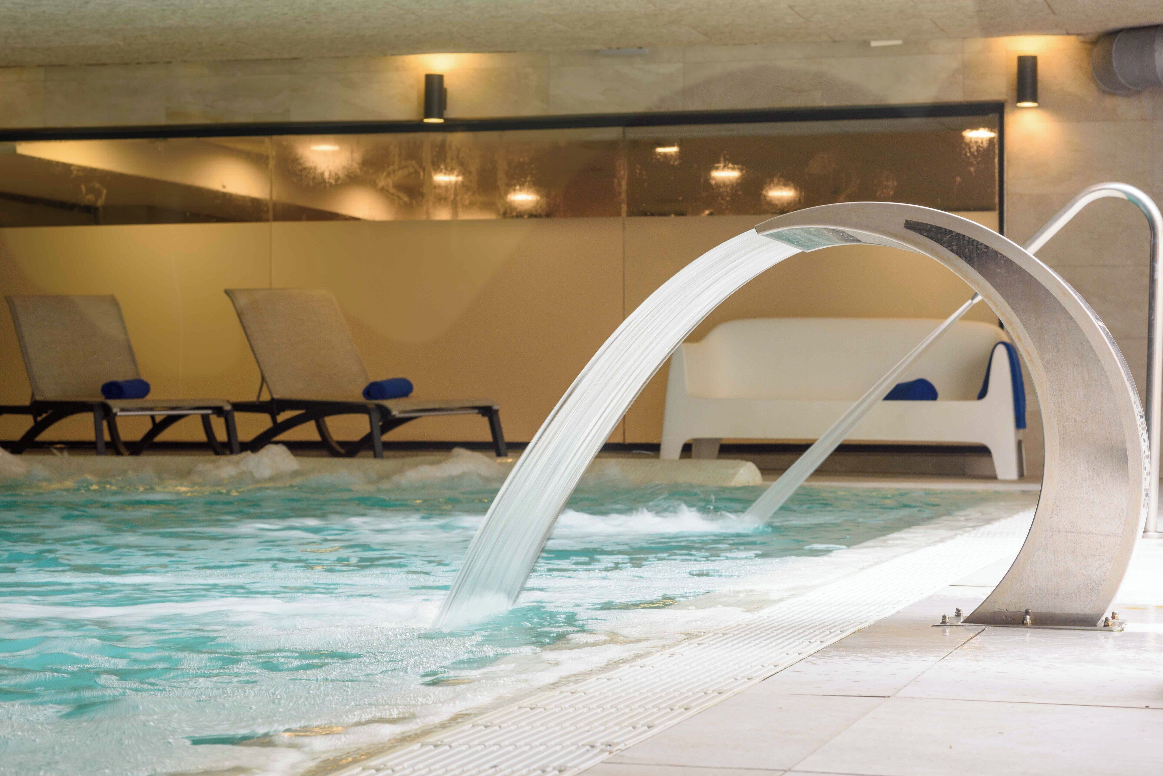 Una piscina interior con agua cristalina, un elemento acuático y sillones de descanso a un lado.