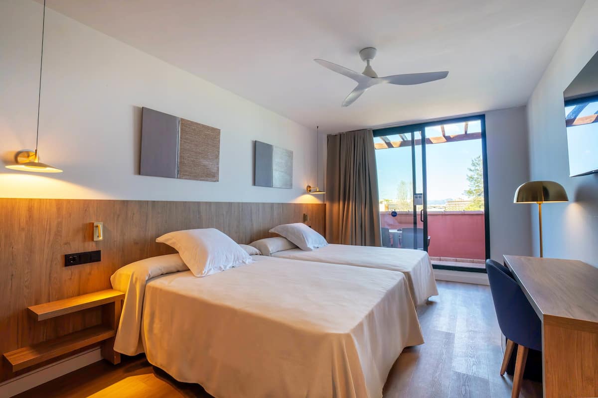 Een hotelkamer met twee bedden, een bureau, een plafondventilator en een groot raam met gordijnen.