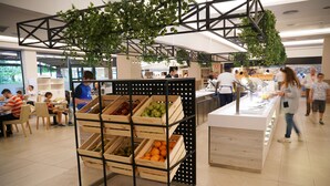 Een moderne, overdekte ruimte met een centraal vertoonde presentatie van houten kisten met fruit en groenten, omringd door zithoeken en een balie waar iemand bediening verleent.