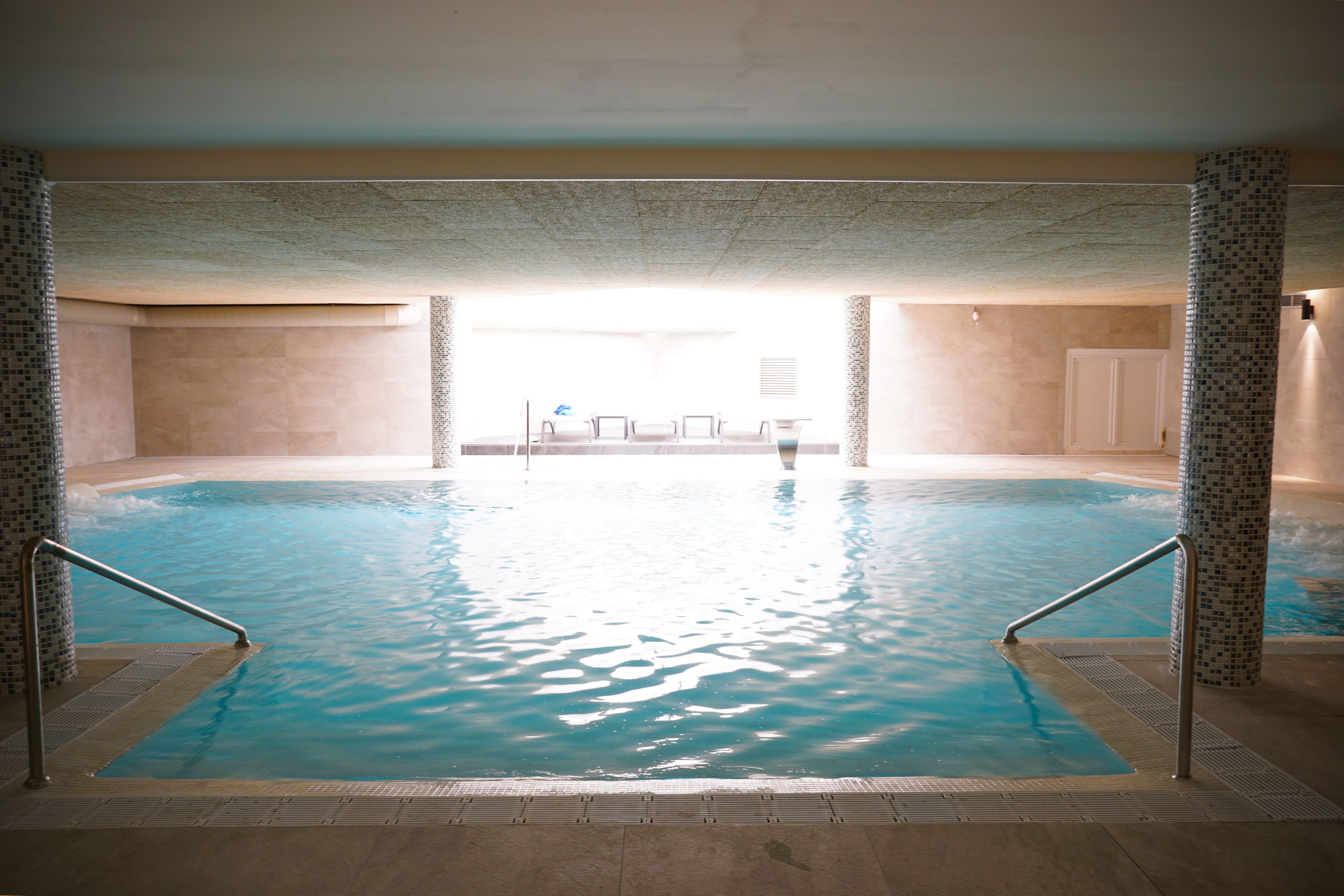 An indoor swimming pool with clear water, tiled walls, and a view of a room with chairs and a table in the background.