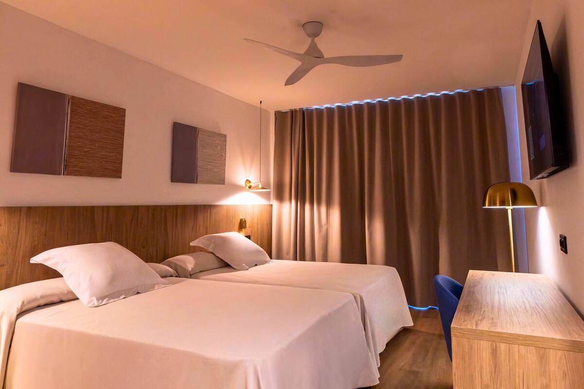 Een hotelkamer met twee bedden, een plafondventilator, een aan de muur bevestigde televisie en een bureau met lamp.