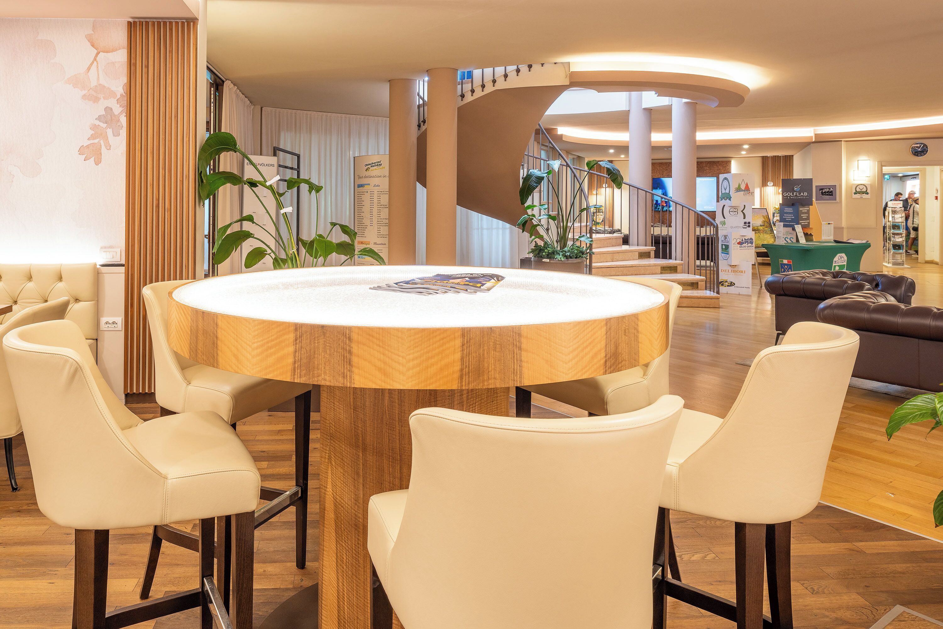 A modern hotel lobby with a round wooden table surrounded by white chairs, a staircase, and a seating area with a brown sofa.