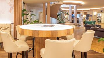 A modern hotel lobby with a round wooden table surrounded by white chairs, a staircase, and a seating area with a brown sofa.