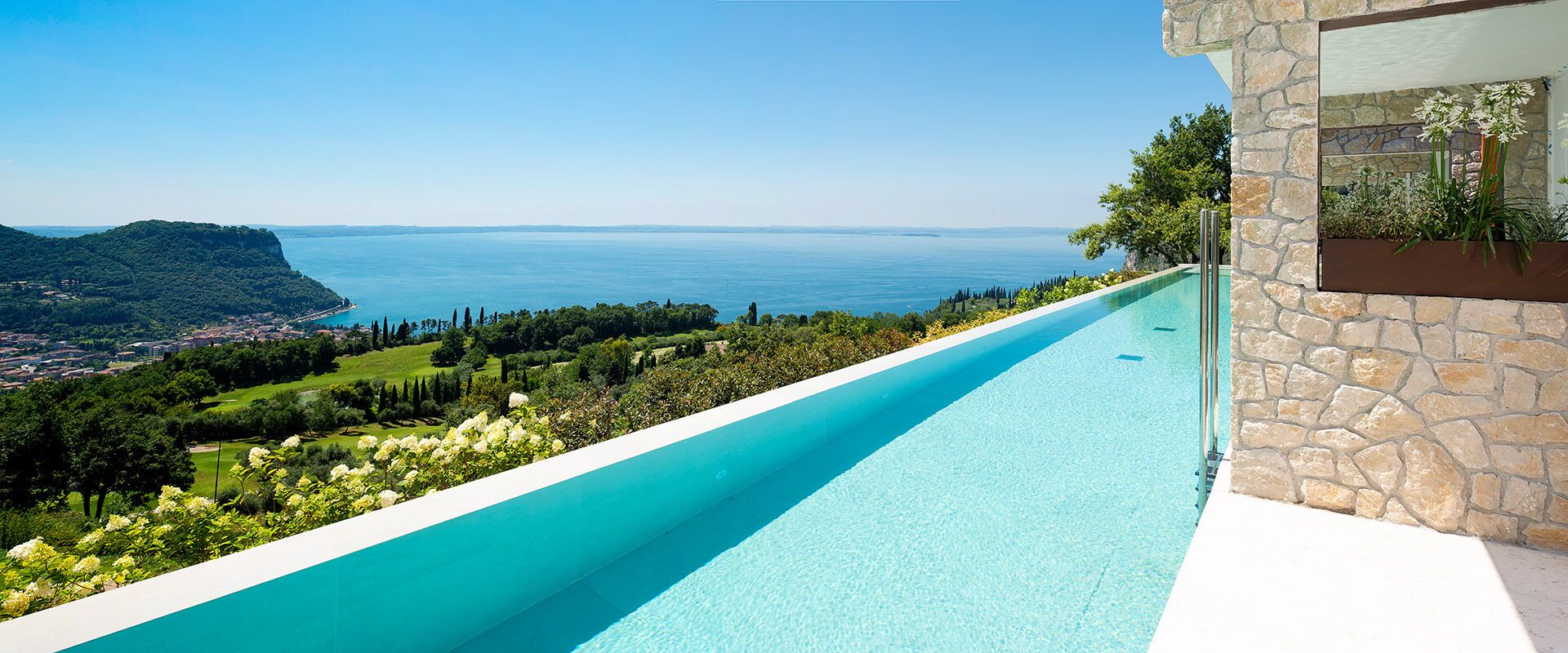 Una piscina larga y estrecha con agua cristalina azul, rodeada de exuberante vegetación y un muro de piedra.