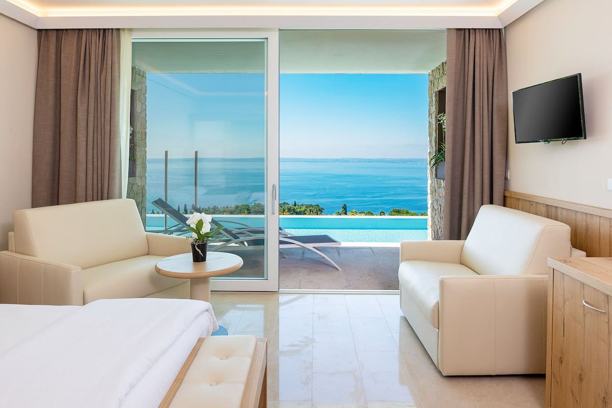 Une chambre d’hôtel dotée d’une grande porte-fenêtre coulissante donnant sur un balcon avec vue sur la mer. On y trouve deux canapés blancs, une petite table ronde et un lit recouvert d’une couette blanche.