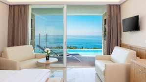 A hotel room with a large sliding glass door leading to a balcony with a sea view. There are two white sofas, a small round table, and a bed with a white duvet.