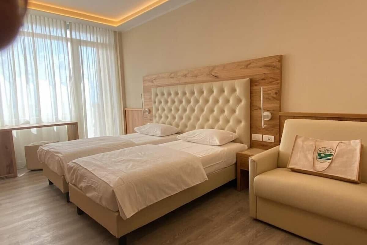 Une chambre d’hôtel avec deux lits, une tête de lit en bois, un canapé et une fenêtre avec des rideaux.