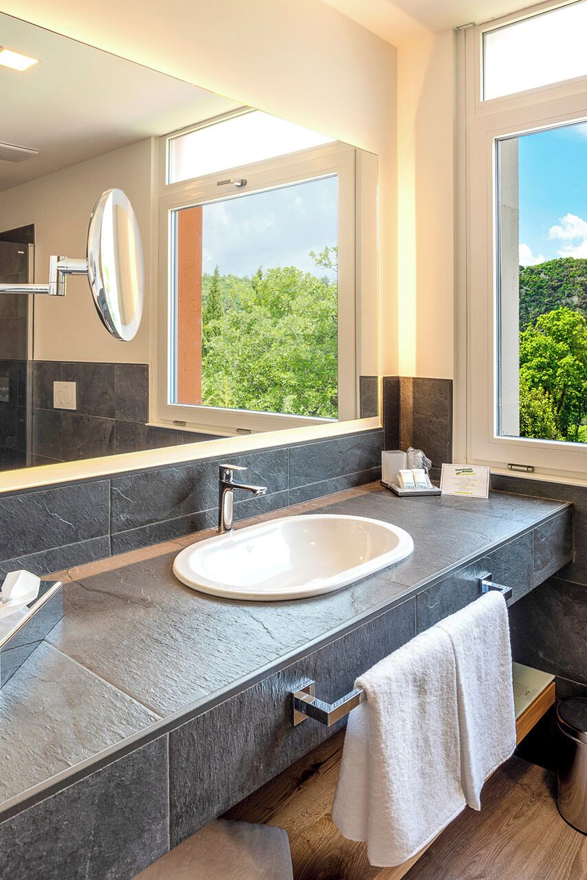 A modern bathroom with a large mirror, a bathtub, and a window offering a view of greenery.