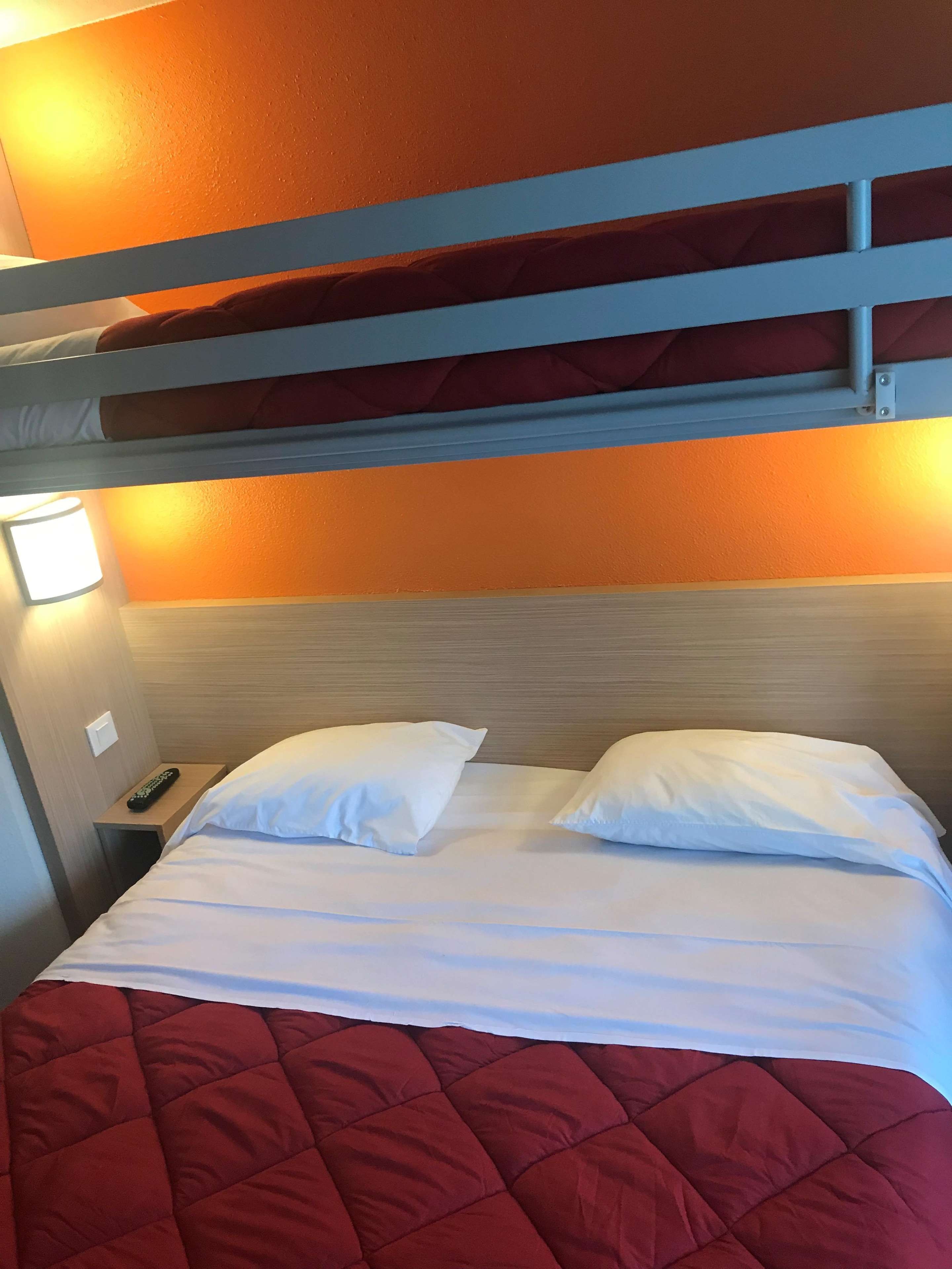 A bunk bed with a red quilt, a bedside lamp, and a remote control on the nightstand.