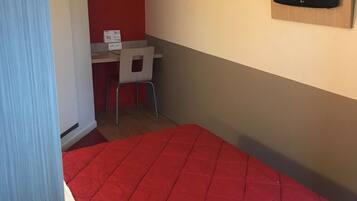 Une petite pièce avec un lit rouge, un bureau avec une chaise et un téléviseur fixé au mur.