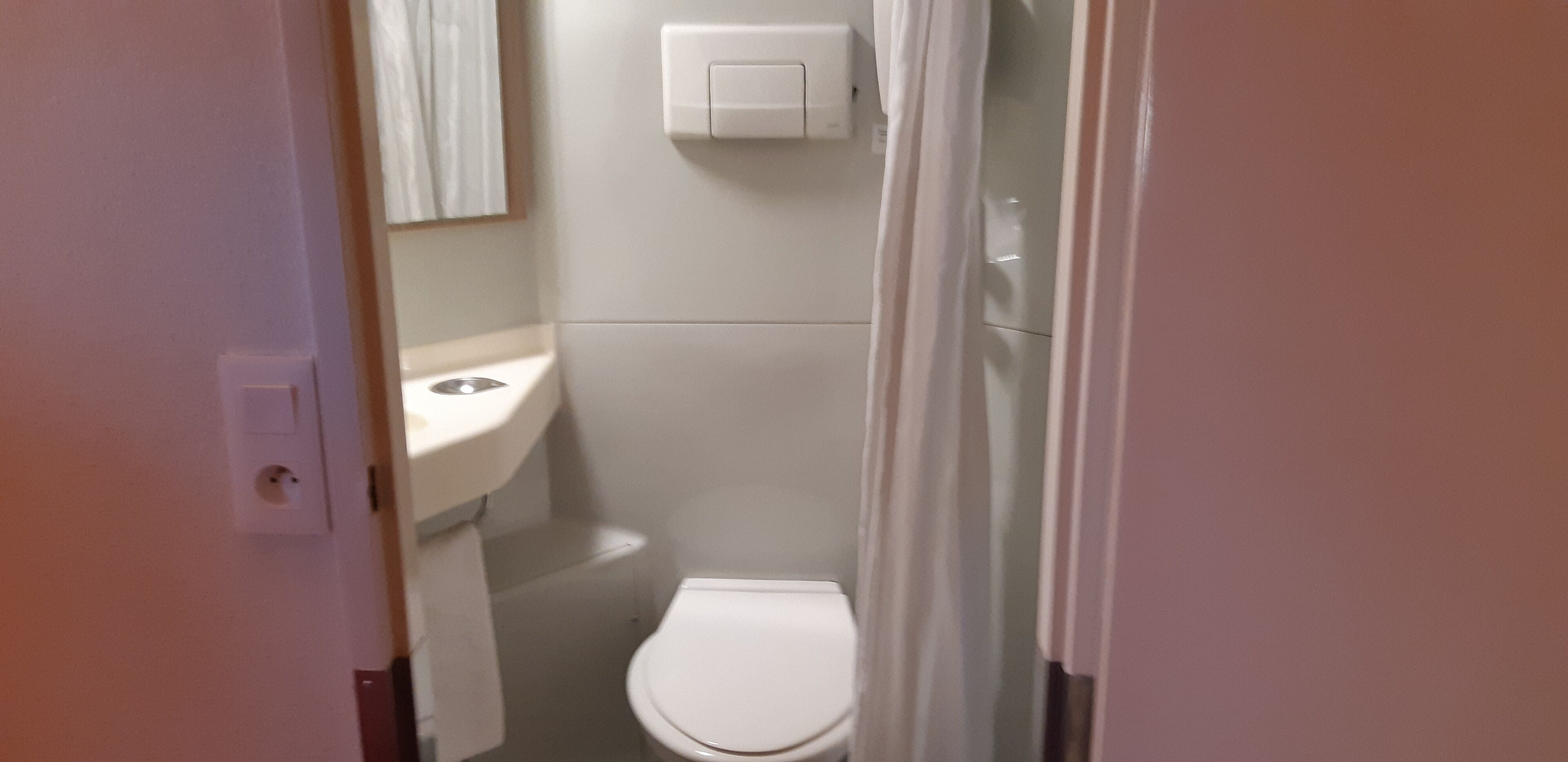 Un baño pequeño y estrecho con inodoro, lavamanos y cortina de ducha, todo en color blanco.