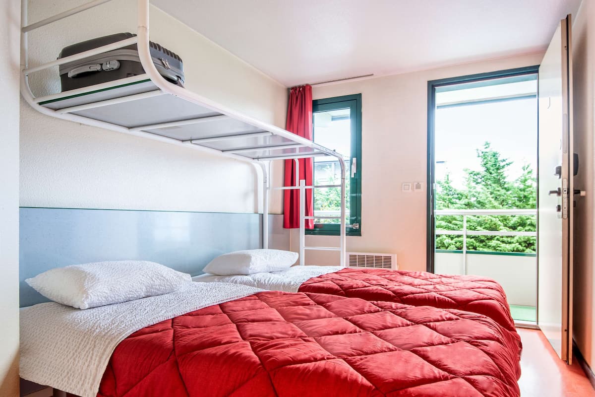 Une chambre avec un lit superposé, une couette rouge matelassée, des oreillers blancs et une couverture blanche. Il y a une fenêtre avec un rideau rouge et une vue sur la verdure à l’extérieur.