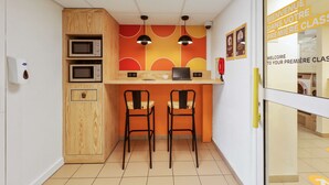 Una kitchenette con dos microondas, un fregadero y un pequeño armario. Hay dos sillas y una mesa con un ordenador portátil. Un letrero dice 'Bienvenue dans votre premiere classe'.