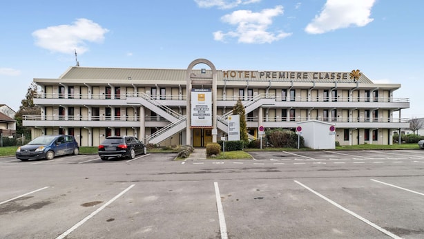 Un hotel de dos pisos, con una entrada central y escaleras, y un estacionamiento con varios coches.